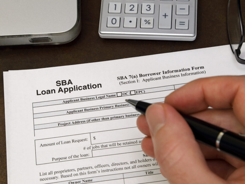 A Small Business Administration aka SBA loan application form, issued by the U.S.A. government, is shown up close with a person holding a pen as if to fill out the application.